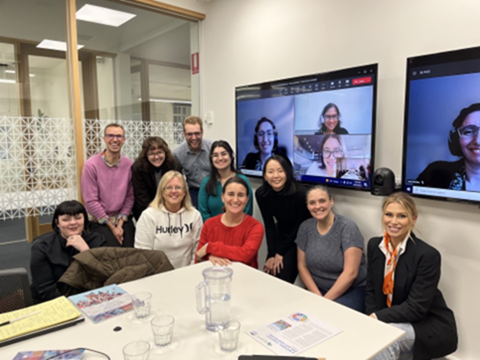 Ten people gather around a table and smile for the camera with three others visible on a Zoom screen behind them.
