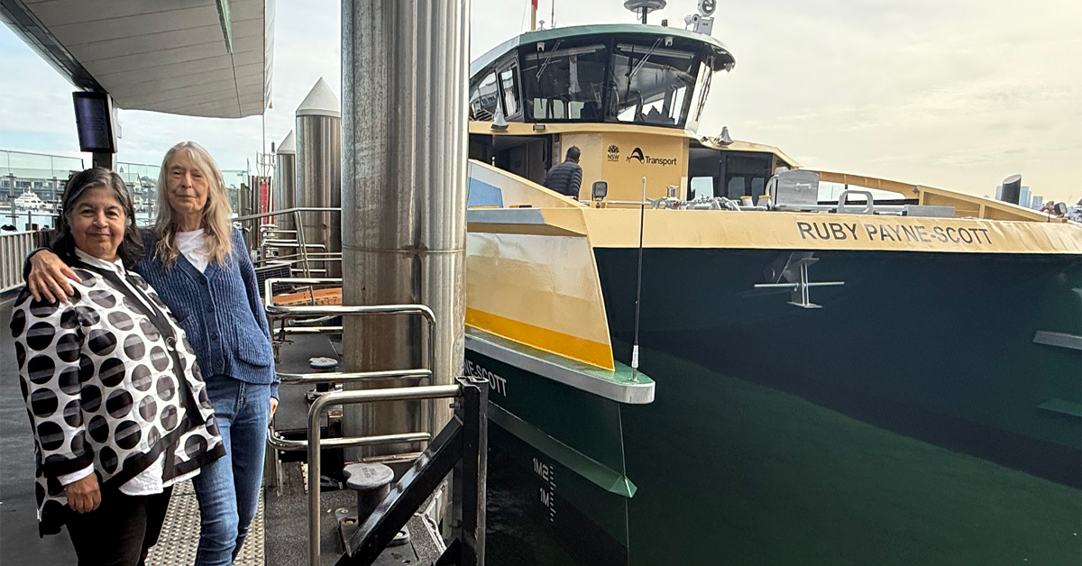 Two women standing on a wharf, next to a green and yellow catamaran ferry named 'Ruby Payne-Scott'