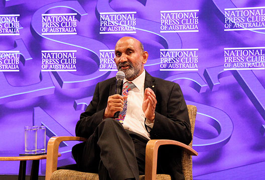 Professor Jagadish sits in a chair speaking into a microphone in front of a purple backdrop which reads 'National Press Club of Australia'