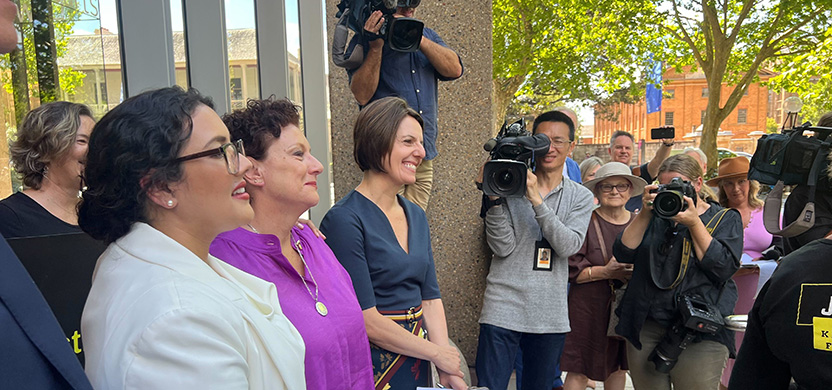 Three women smiling standing in front of a court building surrounded by people with cameras.