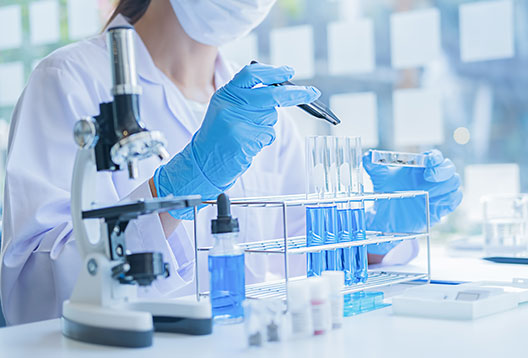 A woman wearing a white lab coat, face mask and blue latex gloves working with test tubes, a petri dish and a microscope